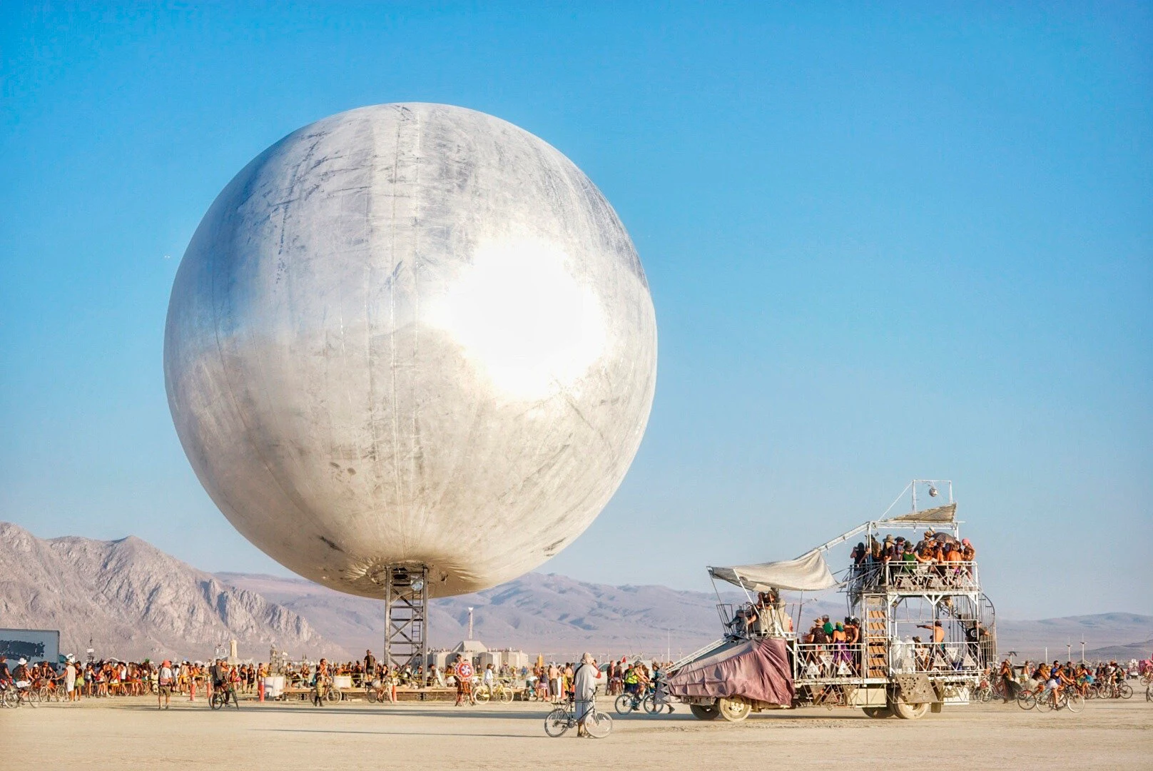 The Orb’s reflective sphere above its supporting mast, surrounded by visitors at Burning Man.
