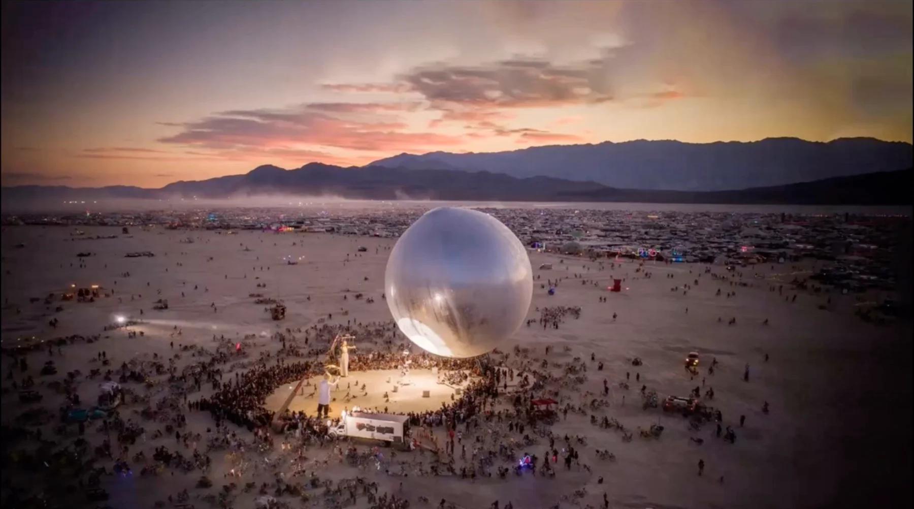 Aerial evening view of The Orb above a crowd at Burning Man, with Black Rock City and mountains beneath a sunset sky.