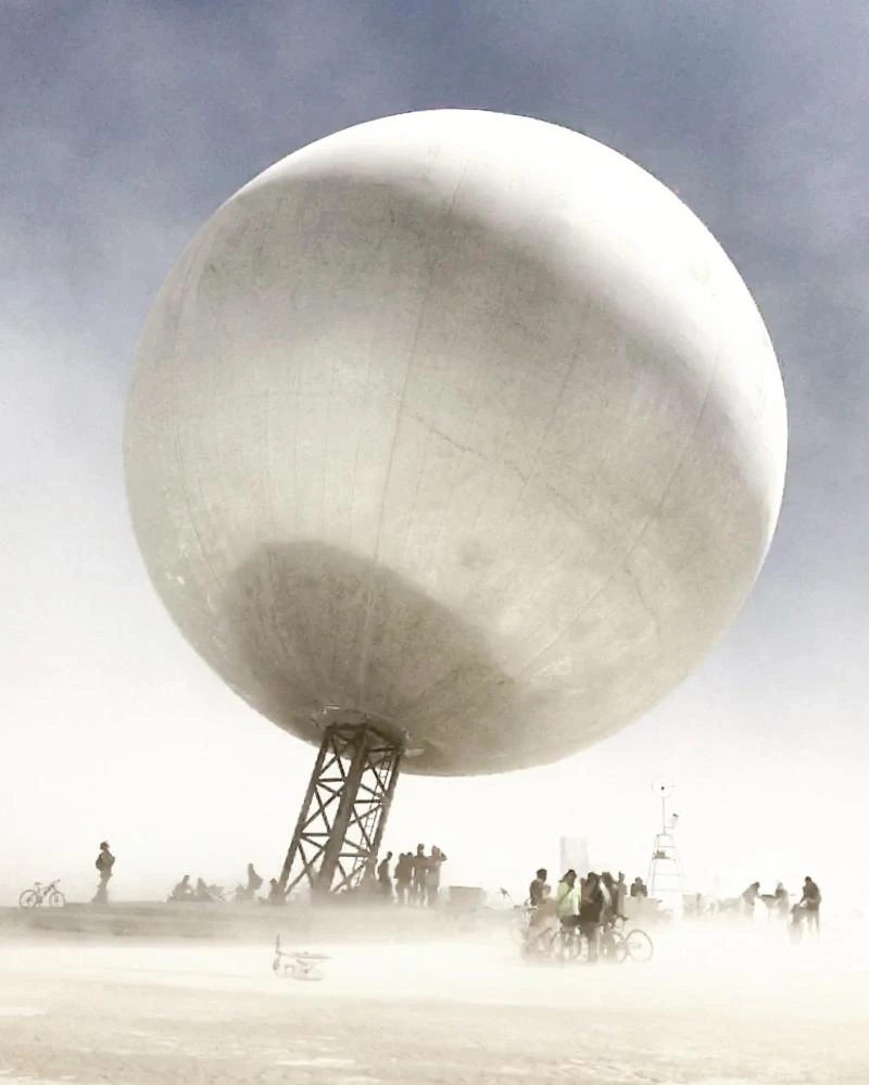 The Orb’s reflective sphere above its inclined lattice mast, with people and bicycles below in the desert dust.